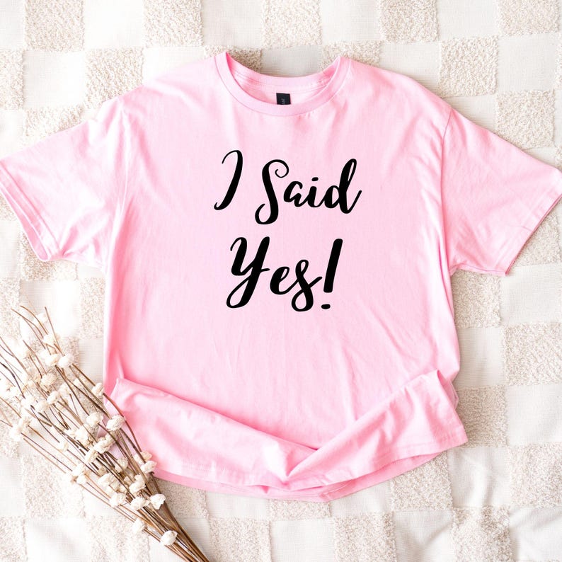 K&ouml;nnte beinhalten: Ein hellrosa T-Shirt mit dem Schriftzug "I Said Yes!" in schwarzer Schrift. Das kurz&auml;rmelige Shirt ist gefaltet, mit einem Zweig wei&szlig;er Blumen in der unteren linken Ecke. Das Shirt liegt auf einer strukturierten, cremefarbenen Oberfl&auml;che.