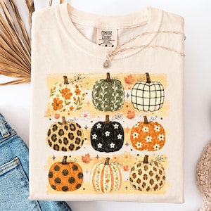 以下が含まれることがあります： クリーム色の半袖Tシャツ。花柄、ヒョウ柄、チェック柄、白い花が描かれた黒いカボチャなど、9種類の異なるパターンのカボチャがグラフィックプリントされており、小さな秋の葉や星のモチーフがアクセントになっています。