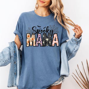 May include: A blue t-shirt with the words "Spooky MAMA" in a Halloween-themed design. The word "Spooky" is in black script with bats and spiderwebs. The word "MAMA" is in a block letter style with Halloween-themed patterns.