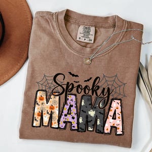 May include: A brown Comfort Colors t-shirt with the words "Spooky Mama" in a Halloween-themed design. The letters are filled with pumpkins, ghosts, and bats. A silver necklace with a pendant rests on the shirt.
