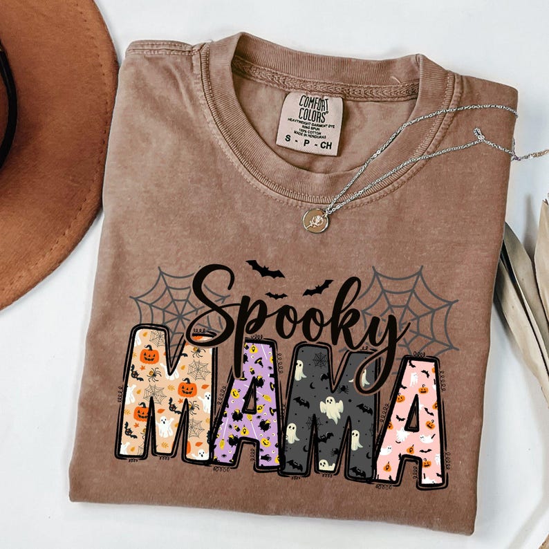 May include: A brown Comfort Colors t-shirt with the words "Spooky Mama" in a Halloween-themed design. The letters are filled with pumpkins, ghosts, and bats. A silver necklace with a pendant rests on the shirt.