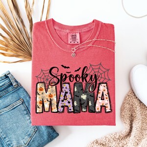 May include: A red t-shirt with the words "Spooky Mama" in a Halloween-themed design. The letters are filled with patterns of pumpkins, ghosts, and bats. The shirt is folded, with a silver necklace and a pair of jeans visible.