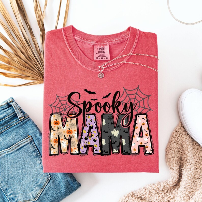 May include: A red t-shirt with the words "Spooky Mama" in a Halloween-themed design. The letters are filled with patterns of pumpkins, ghosts, and bats. The shirt is folded, with a silver necklace and a pair of jeans visible.