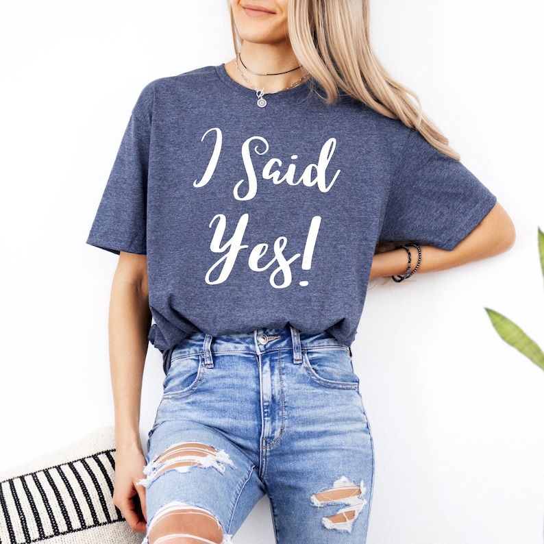 K&ouml;nnte beinhalten: Ein marineblaues T-Shirt mit dem wei&szlig;en Schriftzug "I Said Yes!". Das Shirt wird mit hellblauen zerrissenen Jeans kombiniert. Der Look ist l&auml;ssig und entspannt.