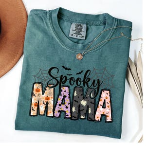 May include: A teal Comfort Colors t-shirt with the words "Spooky Mama" in a Halloween-themed design. The letters are filled with pumpkins, ghosts, and bats. A gold necklace is draped across the shirt.