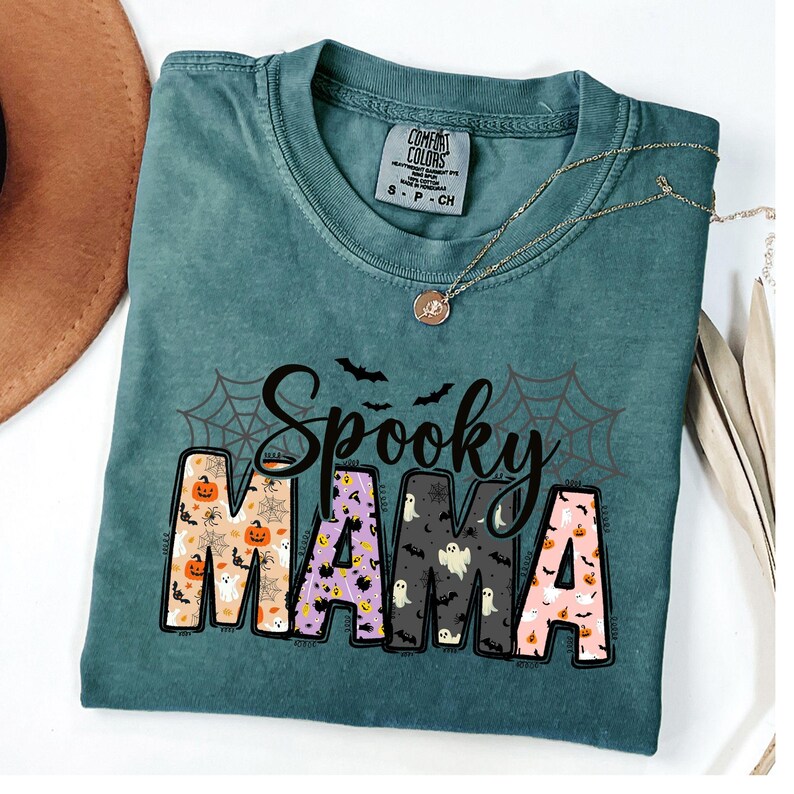 May include: A teal Comfort Colors t-shirt with the words "Spooky Mama" in a Halloween-themed design. The letters are filled with pumpkins, ghosts, and bats. A gold necklace is draped across the shirt.