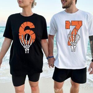 May include: Black and white t-shirts with orange numbers 6 and 7, each featuring a skeleton hand graphic. The shirts are worn by two people standing on a beach. The number 6 is on a black shirt, and the number 7 is on a white shirt.