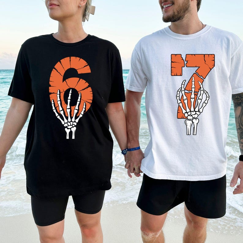 May include: Black and white t-shirts with orange numbers 6 and 7, each featuring a skeleton hand graphic. The shirts are worn by two people standing on a beach. The number 6 is on a black shirt, and the number 7 is on a white shirt.
