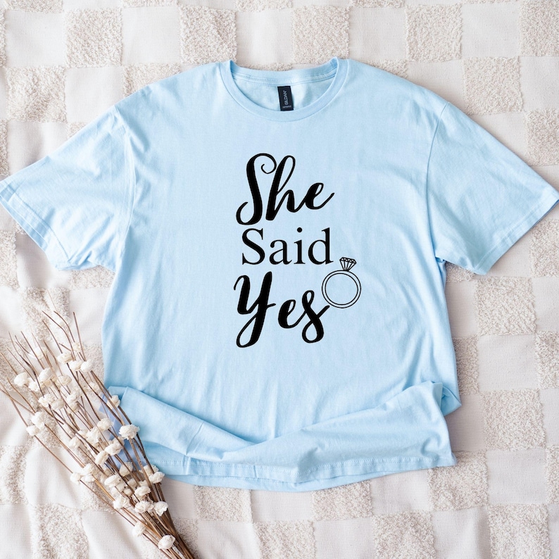 K&ouml;nnte beinhalten: Hellblaues T-Shirt mit dem Text "She Said Yes" in schwarzer Schrift, erg&auml;nzt durch eine Ringgrafik. Das Shirt besteht aus einem weichen Material und ist wahrscheinlich ein Artikel zum Thema Hochzeit oder Verlobung.