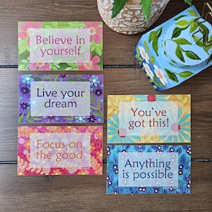 May include: A collection of rectangular cards with floral designs and inspirational quotes. The cards feature phrases like "Believe in yourself," "Live your dream," and "You've got this!" The color palette includes pink, purple, and blue.