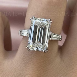 May include: A silver ring featuring a large, rectangular emerald-cut diamond. Two smaller, rectangular diamonds are set either side of the main stone. The ring is on a finger, with a soft pink backdrop.