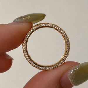 May include: A gold ring featuring a faceted, geometric band design. The outer surface of the ring is lined with a continuous row of small, clear, sparkling stones set into the metal, creating a bright, reflective, and textured appearance.