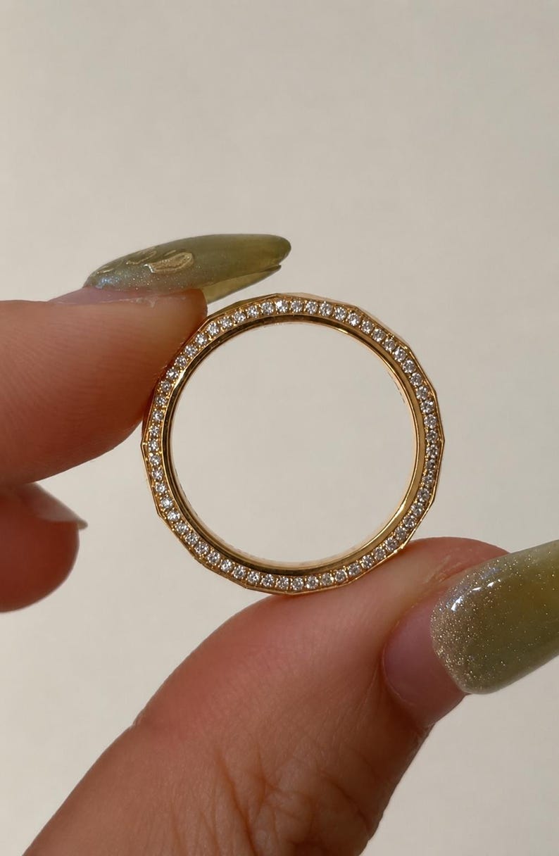 May include: A gold ring featuring a faceted, geometric band design. The outer surface of the ring is lined with a continuous row of small, clear, sparkling stones set into the metal, creating a bright, reflective, and textured appearance.