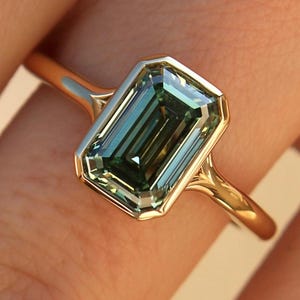 May include: A gold ring with an emerald-cut teal gemstone. The gemstone is set within a gold bezel, and the band is polished gold. The ring is displayed on a finger.