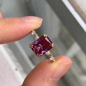 Könnte beinhalten: Ein goldener Ring mit einem großen, rechteckigen, tiefroten Edelstein mit violetten Untertönen. Der Ring hat kleine, klare Akzentsteine und ein zartes Band. Der Ring wird zwischen zwei Fingern gehalten.