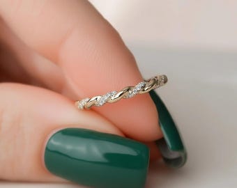 Puede incluir: Un anillo dorado con un diseño de banda trenzada, adornado con pequeñas piedras preciosas transparentes. El anillo se sostiene entre los dedos con esmalte de uñas verde oscuro.