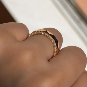 May include: A rose gold ring featuring a faceted geometric outer band and a thin inner band encrusted with a row of small, clear, sparkling round-cut stones. The metal has a polished finish that reflects light across its angular surfaces.