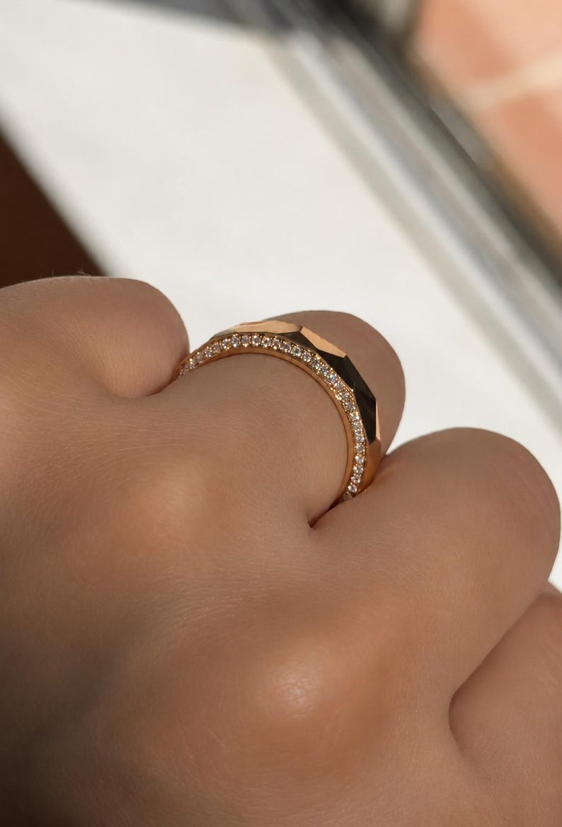 May include: A rose gold ring featuring a faceted geometric outer band and a thin inner band encrusted with a row of small, clear, sparkling round-cut stones. The metal has a polished finish that reflects light across its angular surfaces.
