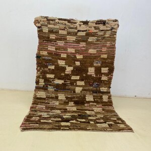 May include: A rectangular, woven rug with a geometric pattern in shades of brown, beige, and pink. The rug has a textured surface and a folded shape, showcasing its design and craftsmanship. The rug is made of natural fibers.