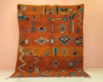 Puede incluir: Una vibrante alfombra tejida a mano con un fondo naranja texturizado. La alfombra presenta patrones geométricos en tonos de azul, blanco y marrón. El diseño incluye formas de diamante, triángulos y motivos abstractos.