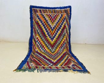 May include: A colorful, rectangular rug with a geometric pattern. The rug features a vibrant array of colors including blue, red, yellow, and orange. The border is a deep blue, and the rug has a fringed edge.