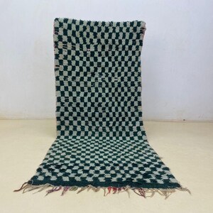 May include: A rectangular, hand-woven rug with a checkerboard pattern in dark green and off-white. The rug has a shaggy texture and a fringed edge. The rug is displayed against a plain white wall and floor.