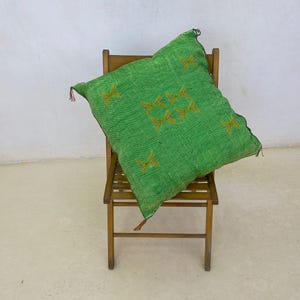 May include: A green decorative pillow with embroidered gold-colored starburst patterns rests on a wooden chair. The pillow has a textured appearance and tassels on the corners. The chair is made of wood and has a simple design.