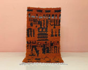 Puede incluir: Una alfombra rectangular tejida a mano con un fondo naranja texturizado y patrones geométricos negros. La alfombra tiene una franja gris a lo largo del borde inferior. La alfombra es un revestimiento decorativo para el suelo.
