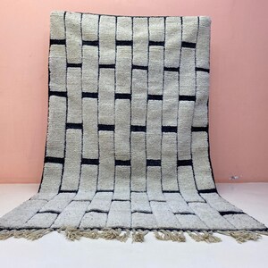Peut inclure: Tapis rectangulaire avec un motif tissé de rectangles blanc cassé et bleu foncé. Le tapis a une frange le long du bord inférieur. Le tapis est présenté contre un mur rose et une surface blanche.