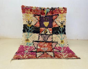 May include: A colorful, handmade Moroccan rug with a geometric design. The rug features a mix of pink, red, black, yellow, and cream colors. The rug is made of a thick, textured material and is likely used as a floor covering.