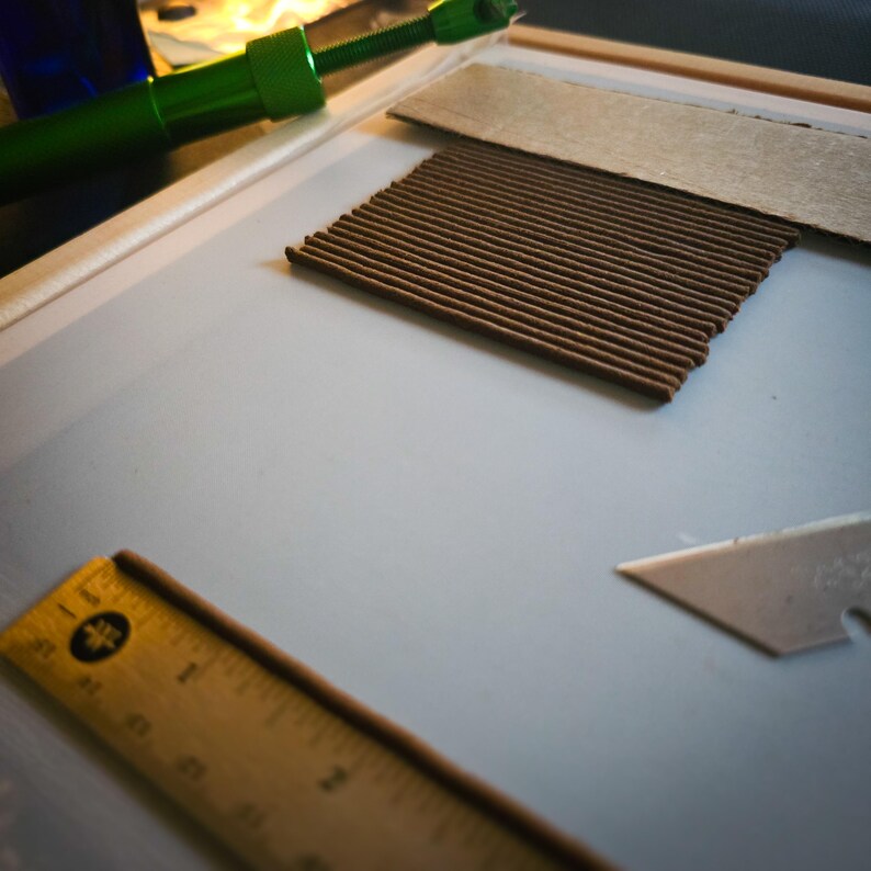 May include: An overhead shot of craft supplies on a white surface. A green tool, a brown corrugated material, a wooden ruler, and a utility knife are visible. The ruler is marked with black numbers and lines.