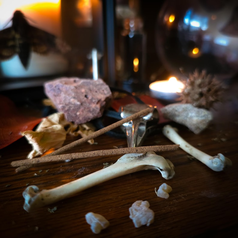 May include: A collection of natural and ritualistic items, including incense sticks, stones, and bones, arranged on a wooden surface. The scene is dimly lit, with a candle and other objects in the background, creating a mystical atmosphere.
