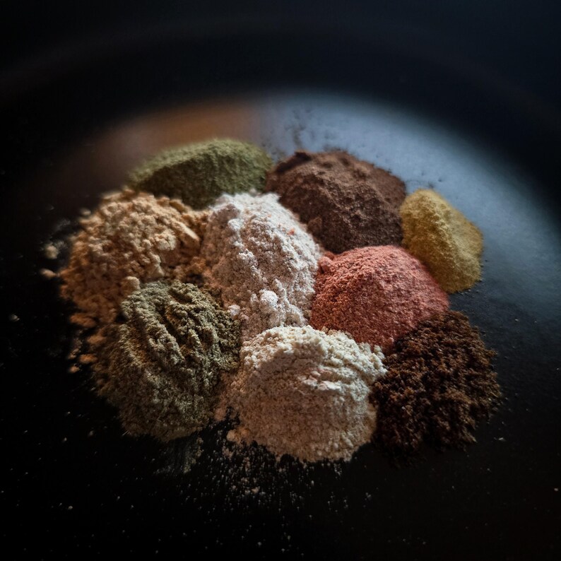 May include: A collection of various ground spices, including shades of green, brown, and red, arranged in a circular pattern on a dark surface. The spices appear finely ground and textured, suggesting a culinary theme.