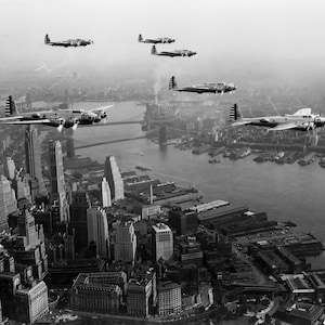 May include: A monochrome aerial view showing a formation of B-17 Flying Fortress aircraft flying above a city. The image captures the planes in flight over a cityscape with a river and buildings. The photograph is taken from a high vantage point.