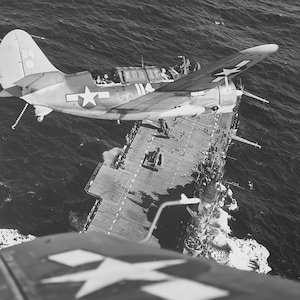 May include: Black and white aerial photograph of a military aircraft approaching an aircraft carrier at sea. The plane has a white star on its wing and the number 114 on its tail. The carrier is filled with people and equipment.