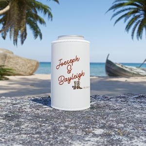 May include: White can cooler with the names "Joeseph & Bayleigh" in brown script, along with a pair of cowboy boots and the date "10.19.24". The background shows a beach scene with palm trees and a boat.