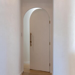 Puede incluir: Una puerta blanca arqueada, ligeramente entreabierta, con un tirador de lat&oacute;n. Revela una habitaci&oacute;n luminosa con suelo de madera. Las paredes blancas crean un espacio interior brillante y aireado. El dise&ntilde;o sugiere una est&eacute;tica moderna.