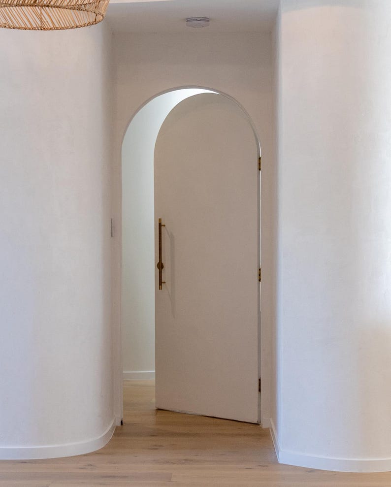 Puede incluir: Una puerta blanca arqueada, ligeramente entreabierta, con un tirador de lat&oacute;n. Revela una habitaci&oacute;n luminosa con suelo de madera. Las paredes blancas crean un espacio interior brillante y aireado. El dise&ntilde;o sugiere una est&eacute;tica moderna.