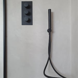 Puede incluir: Un juego de ducha negro mate contra una pared gris claro. El juego incluye una placa rectangular con tres perillas de control redondas y un cabezal de ducha de mano con una manguera flexible.