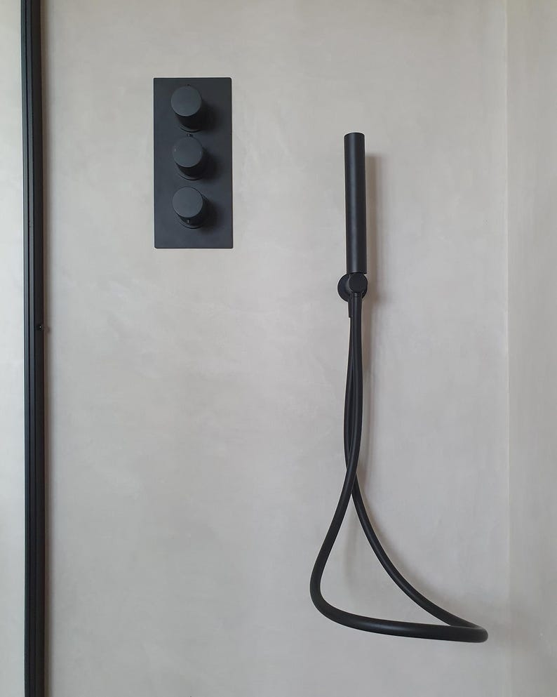 Puede incluir: Un juego de ducha negro mate contra una pared gris claro. El juego incluye una placa rectangular con tres perillas de control redondas y un cabezal de ducha de mano con una manguera flexible.