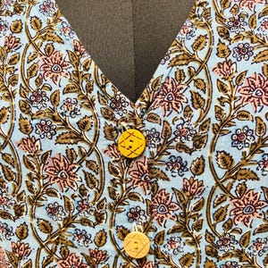 以下が含まれることがあります： ピンク、茶色、白の花柄の、水色の衣服のクローズアップ。 衣服はVネックで、2つの黄色のボタンで留められています。 ボタンは星のようなデザインです。
