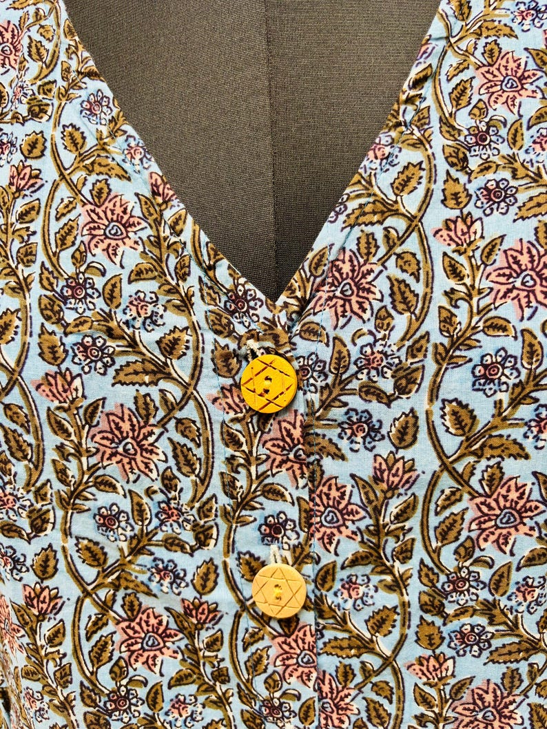 以下が含まれることがあります： ピンク、茶色、白の花柄の、水色の衣服のクローズアップ。 衣服はVネックで、2つの黄色のボタンで留められています。 ボタンは星のようなデザインです。