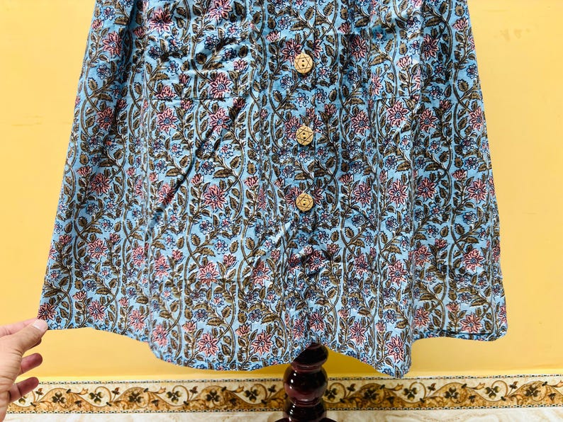 以下が含まれることがあります： ピンクと茶色の花柄の、木製ボタンが付いた水色の衣服。生地には花とツルの詳細なプリントが施されています。この衣服はおそらくトップスまたはドレスです。