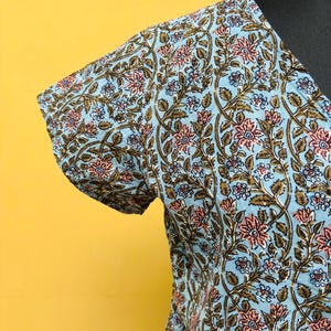以下が含まれることがあります： ライトブルーのフローラルプリントのトップス。生地は、ピンクの花、茶色の葉、小さな青い花がライトブルーの背景に繰り返されるパターンです。トップスは黒いマネキンに表示されています。
