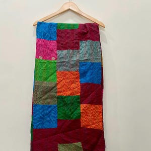 Könnte beinhalten: Ein farbenfroher Patchwork-Quilt mit verschiedenen quadratischen Mustern in Blau-, Rosa-, Grün-, Orange- und Burgundertönen. Der Quilt hängt an einem Holzbügel vor einer schlichten weißen Wand.