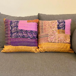 May include: Two decorative throw pillows with patchwork designs. The pillows feature a combination of colors, including purple, pink, and gold. The patchwork design adds a unique and artistic touch to the pillows, perfect for home decor.