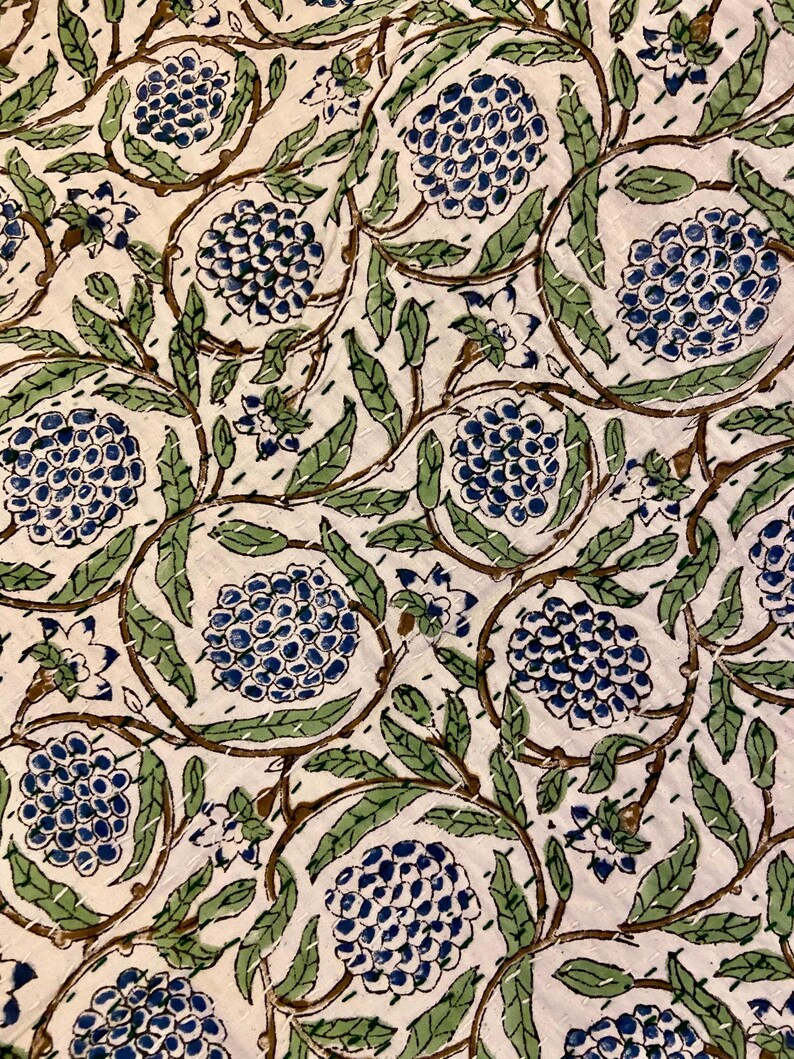 May include: A close-up of a white fabric with a repeating floral pattern. The design features blue, green, and brown elements, including stylized flowers and leaves. The fabric is stitched with white thread, creating a quilted effect.