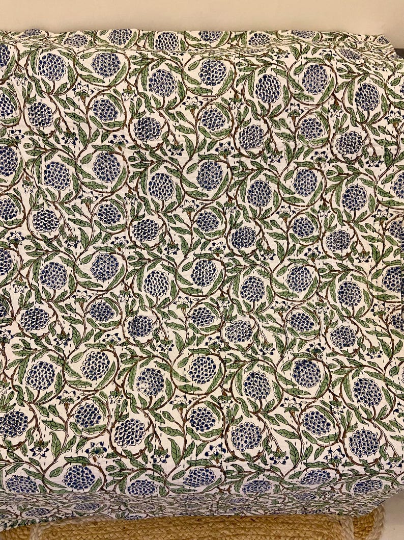 May include: A white fabric with a repeating pattern of blue floral clusters and green leaves connected by brown stems. The design is densely packed, creating a textured appearance. The fabric is likely used for home decor.