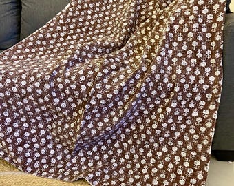 May include: A brown and white floral patterned throw blanket draped over a gray sofa. The blanket features a repeating pattern of white flowers on a brown background. The blanket is made of a soft material and is folded over the sofa.