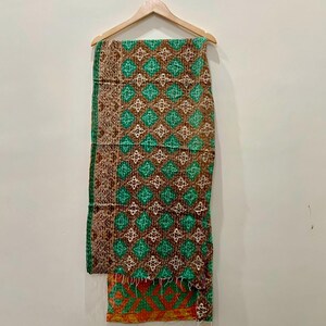 May include: A patterned textile hanging on a wooden hanger. The textile features a repeating diamond pattern in shades of green, brown, and white. The bottom edge has an orange and green geometric design. The textile is likely a blanket or throw.